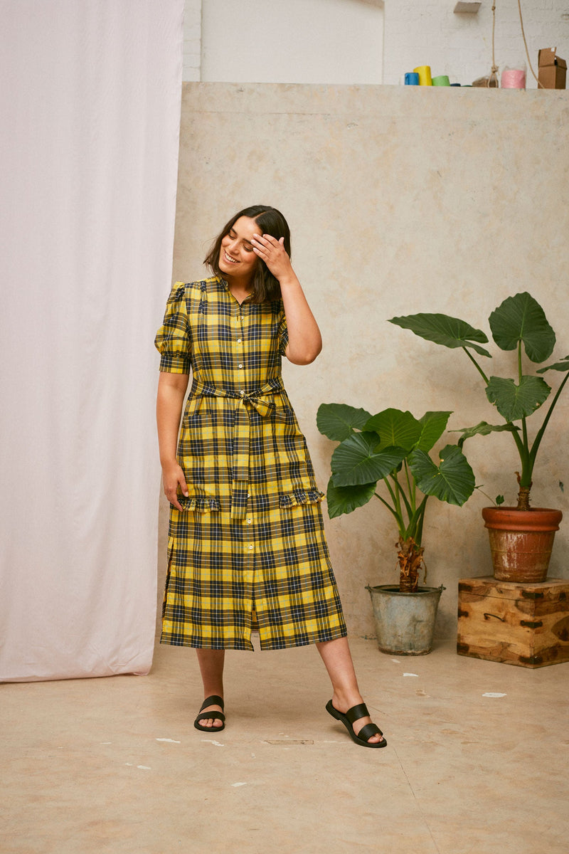 Full length shot of model wearing Saywood's Rosa yellow check shirtdress, with belt loosely tied round the waist. Puff sleeves and patch pockets with ruffles are on the dress. Model has one hand to her head and the dress is worn with black sandals. A plant and drop of pink fabric can be seen in the background.