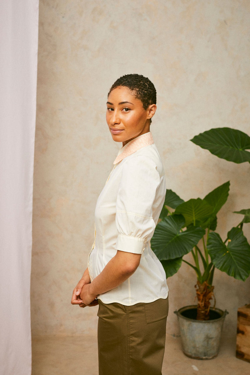 Profile view of model wearing Saywood's pastel yellow Joni puff sleeve blouse with orange lace collar and scalloped hem. Worn with khaki Amelia wide leg trousers. A plant and drop of pink fabric can be seen in the background. 