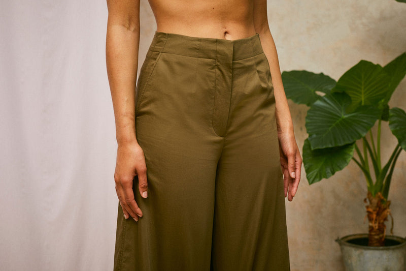 Cropped image of models wearing Saywood's Amelia khaki wide leg trouser, angled front waist view. A plant and drop of pink fabric can be seen in the background.