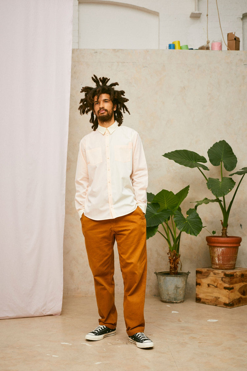 Full length shot of model wearing Saywood's Eddy Mens mens patchwork Shirt with patch pockets in orange and yellow. Worn with tabacco trousers and black Converse. Model has both hands in his trouser pocket and a plant and drop of pink fabric can be seen in the background.