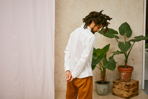 Back view of model wearing Saywood's Eddy Mens mens white Shirt. Worn with tabacco trousers just visible. Model has both hands behind his back and looks towards the floor. A plant and drop of pink fabric can be seen in the background.