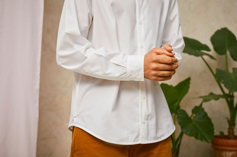 Close up of model's waist, wearing Saywood's Eddy Mens mens white Shirt. Worn with tabacco trousers just visible. Model holds both hands in front of his waist, showing the shirt cuff. A plant and drop of pink fabric can be seen in the background.