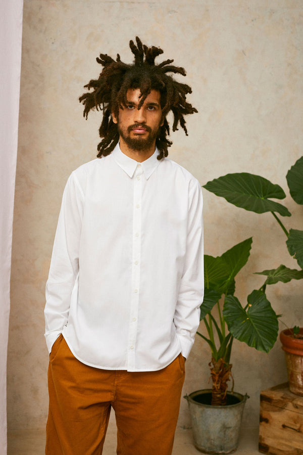 Close up of model wearing Saywood's Eddy Mens mens white Shirt. Worn with tabacco trousers just visible. Model has both hands in his trouser pocket and a plant and drop of pink fabric can be seen in the background.