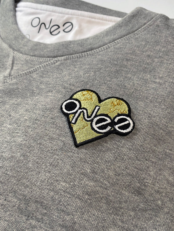 ONE Love embroidered sew on patch, white ONEE lettering of ONE Essentials logo on a pale green heart with the world countries outlined shown on a grey marl organic cotton sweatshirt