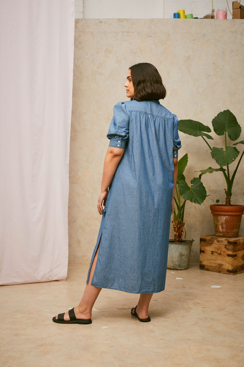 Full length back view shot of model wearing Saywood's Rosa denim shirtdress in light wash Japanese denim; gathers can be seen on the back of the dress from the yoke seam. Worn with black sandals, with one leg out to the side. A plant and drop of pink fabric can be seen in the background.