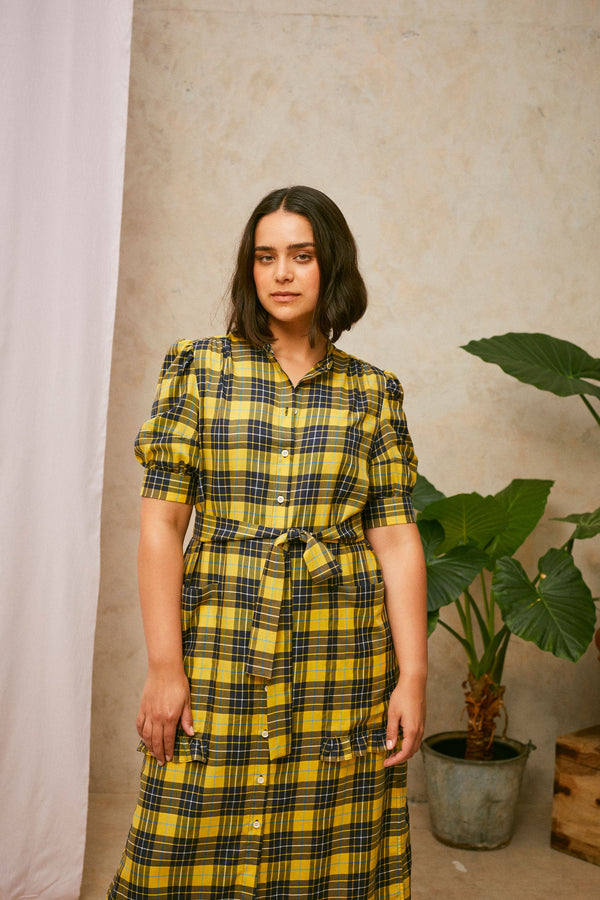 Close up crop of model wearing Saywood's Rosa yellow check shirtdress, with belt loosely tied round the waist. Puff sleeves and patch pockets with ruffles are on the dress. A plant and drop of pink fabric can be seen in the background.