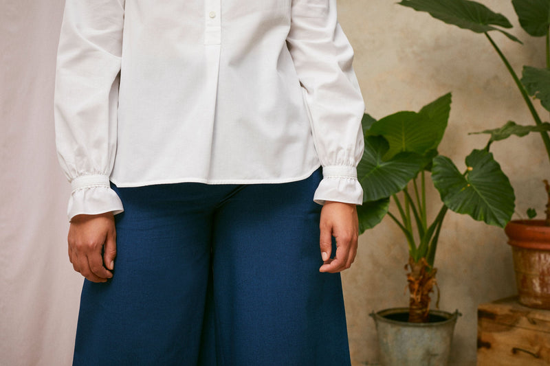 Close up of model wearing Saywood's white shirt, the Marie A-Line Blouse. Her arms are by her sides and the frill cuffs with lace trim are seen. She wears the natural indigo Japanese denim Amelia wide leg trousers. A plant and drop of pink fabric can be seen in the background.