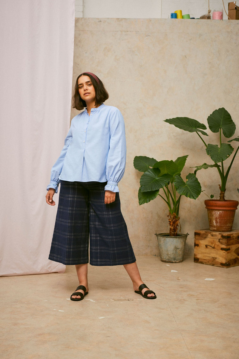 Model stands wearing Saywood's pale blue shirt, the Marie A-Line Blouse, with navy check Amelia wide leg trousers. The red check Heidi Headband is worn in her her. A plant and drop of pink fabric can be seen in the background.