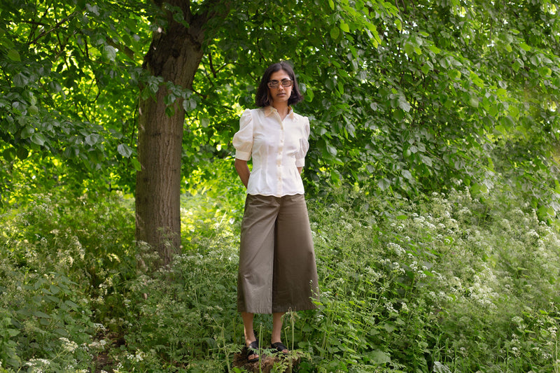 Model stands surrounded by trees and greenery, wearing Saywood's pastel yellow Joni puff sleeve blouse with orange lace collar and scalloped hem. Worn with khaki Amelia wide leg trousers and black sandals and a pair of sunglasses. A plant and drop of pink fabric can be seen in the background. 
