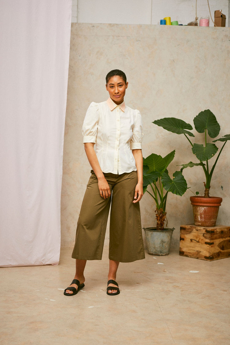 Model stands, wearing Saywood's pastel yellow Joni puff sleeve blouse with orange lace collar and scalloped hem. Worn with khaki Amelia wide leg trousers and black sandals. A plant and drop of pink fabric can be seen in the background. 