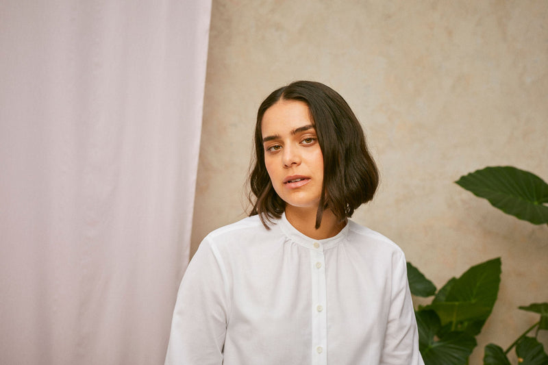 Full length shot of model wearing Saywood's white shirt, the Marie A-Line Blouse. The soft gathers at the neck can be seen. A plant and drop of pink fabric can be seen in the background.