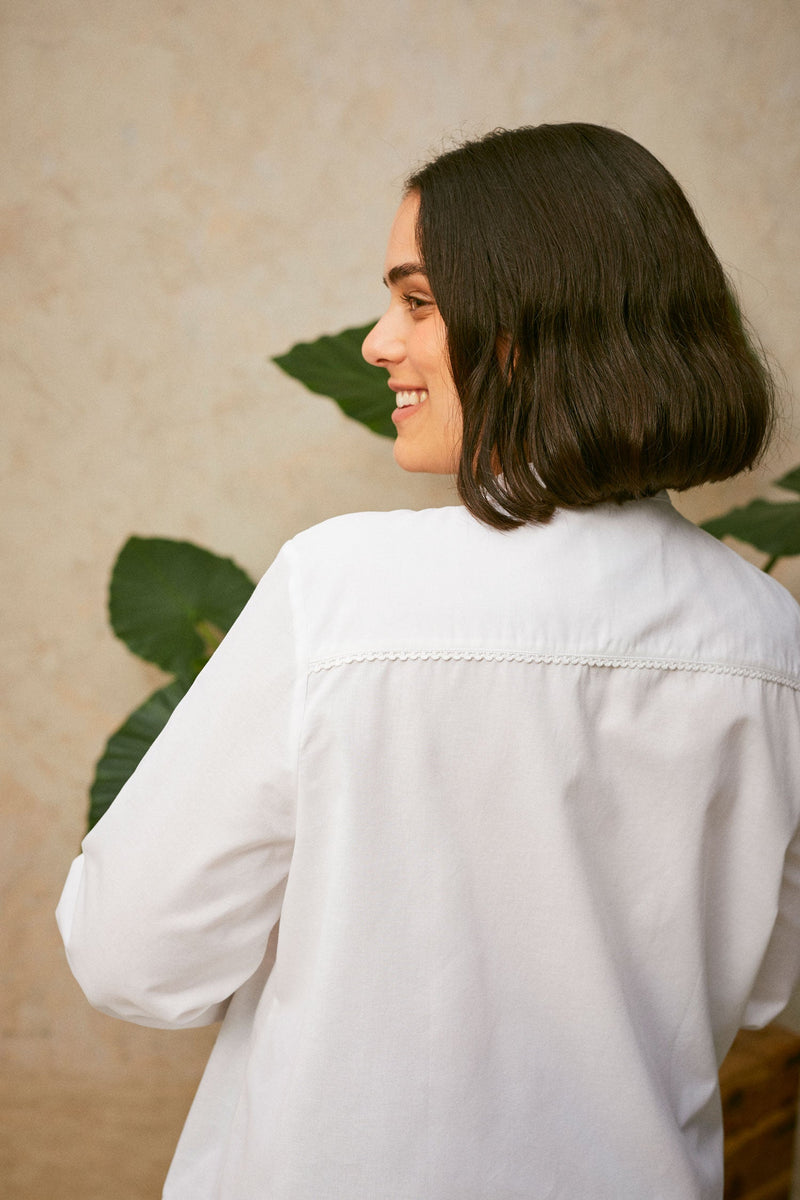 Close up back view of model wearing Saywood's white shirt, the Marie A-Line Blouse. The lace trim at the back neck yoke can be seen. A plant can be seen in the background.