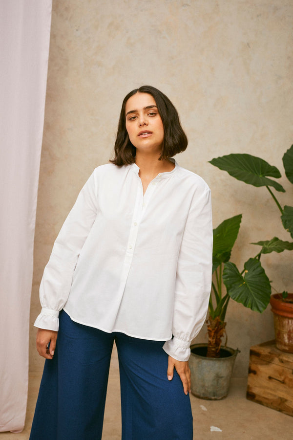 Model wears Saywood's white shirt, the Marie A-Line Blouse, with natural indigo Japanese denim Amelia wide leg trousers. A plant and drop of pink fabric can be seen in the background. 