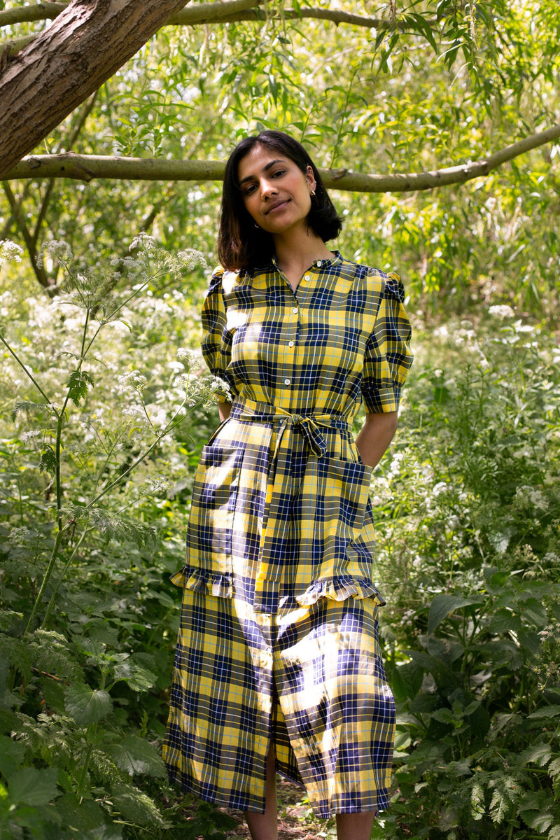 Model stands by a tree surrounded by greenery, wearing Saywood's Rosa yellow check shirtdress, with belt loosely tied round the waist. She smiles and her hands are behind her back, and puff sleeves and patch pockets with ruffles are seen on the dress.