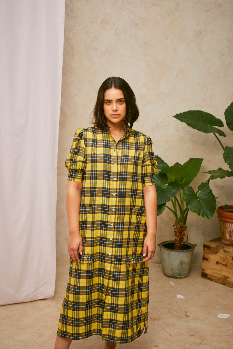 Model is wearing Saywood's Rosa yellow check shirtdress, loosely worn without a belt. Puff sleeves and patch pockets with ruffles are on the dress. A plant and drop of pink fabric can be seen in the background.